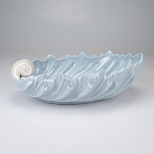 May include: A light blue ceramic bowl shaped like a large leaf with a glossy finish. The bowl features a prominent, white, spiraled shell-like handle on one side and a textured, scalloped edge that mimics the organic veins and curves of a leaf.