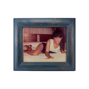 Puede incluir: Fotografía vintage enmarcada con una persona en un traje de baño blanco y azul marino. La foto está dentro de un marco de madera azul desgastado. La persona está acostada en una cama.