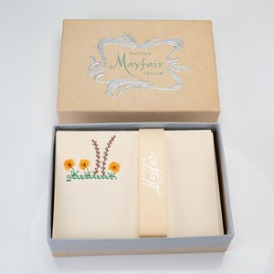 May include: A tan cardboard box labeled Eaton's Mayfair Vellum in silver foil. Inside, a stack of cream-colored stationery is held by a tan paper band with the text Eaton's Mayfair. The top sheet features a small illustration of orange flowers.