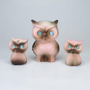May include: A set of three ceramic owl figurines in varying sizes, featuring a soft pink and brown gradient finish. Each owl has two bright blue rhinestone eyes set into the face, with a smooth, rounded body shape and pointed ear tufts.