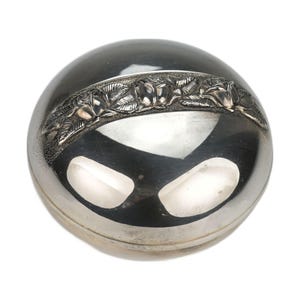 Puede incluir: Un joyero redondo de metal plateado pulido con una banda texturizada en relieve en la parte superior. Esta banda decorativa muestra un patrón detallado de tres rosas en flor rodeadas de hojas sobre un fondo oscuro y contrastado.