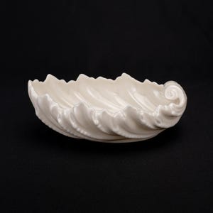 May include: An off-white ceramic bowl shaped like a seashell with a glossy finish. The bowl features a scalloped edge and a spiraled end, mimicking the natural texture and form of a conch shell with deep, curved ridges along the interior surface.