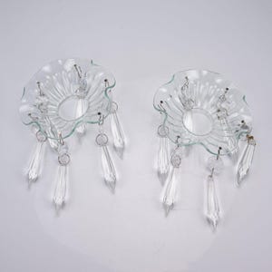 May include: A pair of clear glass candle bobeches featuring scalloped edges and a series of hanging crystal prisms. Each bobeche has a central circular opening and metal rings that secure the faceted, teardrop-shaped glass pendants.
