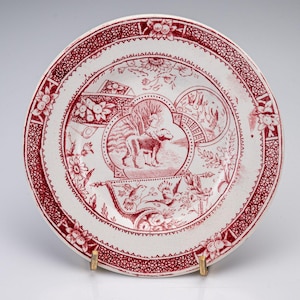 May include: A round ceramic plate featuring a deep red transferware pattern on a white background. The central design depicts a dog standing in a landscape, surrounded by floral motifs, birds, and geometric borders along the scalloped rim.