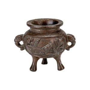 May include: A small, dark bronze-toned metal incense burner featuring a rounded body, three short legs, and two side handles shaped like stylized animal heads. The exterior surface is decorated with embossed floral and bird motifs.