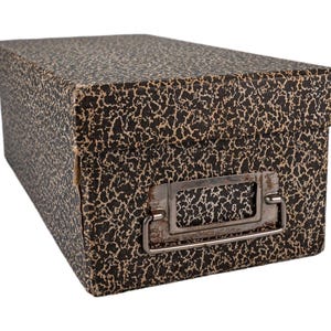 Op de afbeelding: Een rechthoekige opbergdoos met een afneembaar deksel, voorzien van een zwart en beige gespikkeld patroon. De voorzijde van de doos bevat een rechthoekige metalen labelhouder met een scharnierende handgreep, met tekenen van verouderde metaalpatina.