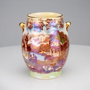以下が含まれることがあります： 虹色の光沢仕上げが施された陶磁器製の花瓶。茶色の転写プリントで城、木々、放牧されている牛の風景が描かれています。花瓶の両側には金色の取っ手があり、縁は金色で、底面は淡い緑色をしています。