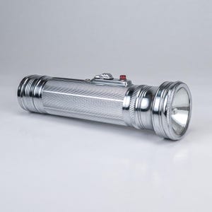 May include: A cylindrical silver-colored metal flashlight with a ribbed handle texture. It features a prominent silver sliding power switch topped with a small red button, a flared head section, and a reflective interior lens housing.