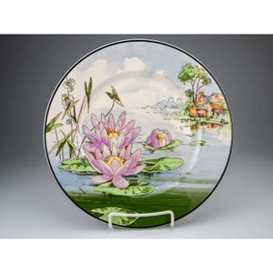 May include: A decorative plate featuring a serene water lily pond scene. The plate showcases pink water lilies with yellow centers, green lily pads, and a lakeside cottage. The plate has a black rim and a white background.