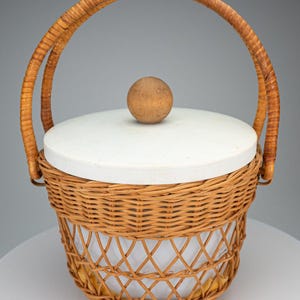 Puede incluir: Una cesta de mimbre vintage con tapa blanca y pomo de madera. La cesta tiene un asa tejida y un diseño de enrejado en la mitad inferior. La cesta es de mimbre marrón claro.