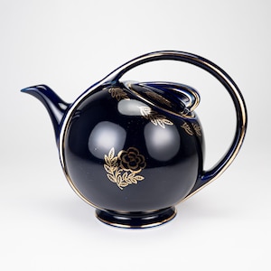 May include: A spherical dark blue teapot featuring a gold floral pattern and gold trim. The teapot has a curved handle that extends from the top to the base, a short spout, and a matching lid with a gold-trimmed edge, all set against a plain background.