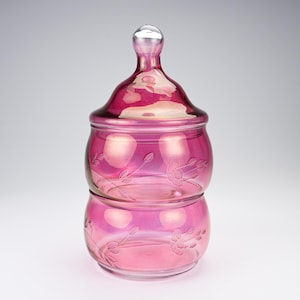 Puede incluir: Un frasco de vidrio magenta de dos niveles con una tapa a juego que presenta un remate redondeado. El vidrio tiene un acabado translúcido y está decorado con patrones florales grabados que incluyen tallos, hojas y flores en el cuerpo.