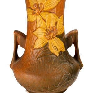 Può includere: Un vaso in ceramica marrone con due manici e una larga apertura. Il vaso presenta fiori gialli in rilievo con foglie verdi. Il vaso ha una superficie strutturata e una base arrotondata. Il design generale ricorda l'Art Nouveau.