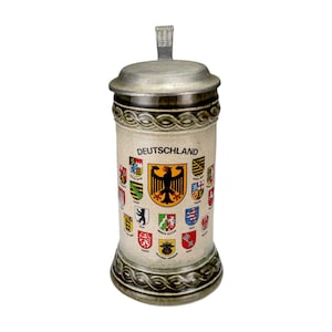 May include: A ceramic beer stein with a hinged metal lid. The stein is beige with the word "Deutschland" and various colorful coat of arms. A black eagle emblem is centered on the stein.