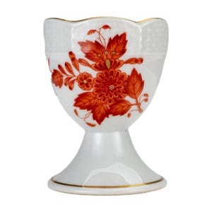 May include: White ceramic egg cup with a flared base and a gold rim. The cup features a red floral design with leaves and a textured upper portion. A decorative piece for serving eggs.