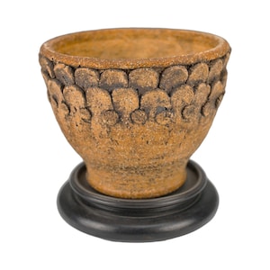 May include: A brown ceramic bowl with a textured, patterned exterior, set on a dark, round base. The bowl's rim and exterior have a detailed, organic design. The overall aesthetic is rustic and earthy, with a handcrafted feel.