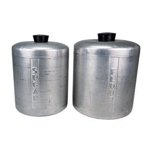 May include: Two vintage aluminum canisters, one labeled "SUGAR" and the other "FLOUR". Each canister has a cylindrical shape with a black lid. The canisters have a brushed metal finish and are likely for kitchen storage.