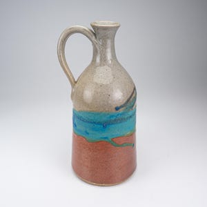 May include: A ceramic pitcher with a tall, slender neck and a single curved handle. The upper portion is a speckled light gray, transitioning into a vibrant horizontal band of bright turquoise blue, and ending in a solid terracotta red base.