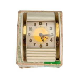 May include: A vintage Westclox clock with a cream-colored face and gold-tone accents. The clock face has black numerals and hands, with a yellow center. The clock is encased in a cream and gold frame, and sits on a cardboard base.