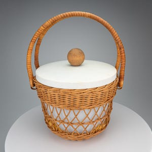 Op de afbeelding: Een ronde gevlochten rieten mand met een witte cilindrische inzet en een bijpassend wit deksel met een kleine houten bolvormige handgreep. De mand heeft een hoog, gebogen, omwikkeld rieten handvat dat aan de zijkanten is bevestigd.