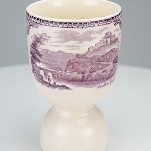 May include: Cream-colored ceramic egg cup with a purple landscape design. The design features a bridge, castle, and trees. The egg cup has a bulbous base and a flared top.