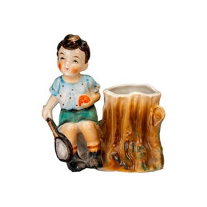 Może przedstawiać: Figurka ceramiczna w stylu vintage przedstawiająca dziecko siedzące obok pnia drzewa. Dziecko ma na sobie niebieską koszulę w białe kropki, zielone szorty i czarne buty. Figurka trzyma rakietę tenisową i czerwony owoc. Pień drzewa ma wydrążony wierzch.
