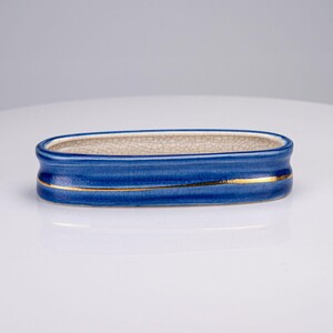May include: An oval-shaped ceramic dish featuring a deep blue glazed exterior with a thin, horizontal gold-colored line accent. The interior of the dish has a light beige crackle glaze finish, showing a fine network of lines across the surface.