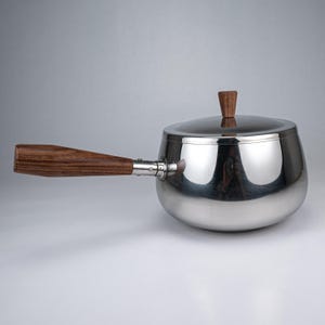 Puede incluir: Un cazo de acero inoxidable pulido con cuerpo redondeado y tapa a juego. El recipiente cuenta con un mango largo y horizontal de madera y un pequeño pomo de madera vertical en el centro de la tapa, ambos con un patrón de veta de madera.