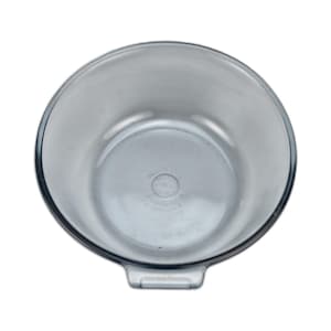 Può includere: Una ciotola rotonda di vetro trasparente con un bordo leggermente svasato. La ciotola ha un interno liscio e una piccola maniglia rettangolare su un lato. Il fondo della ciotola ha un segno circolare. La ciotola è vuota e su sfondo bianco.