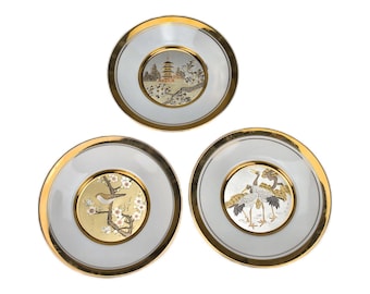 May include: Three circular porcelain plates with gold-toned rims. The top plate features a pagoda landscape. The bottom left plate displays a bird on a flowering branch against a gold background. The bottom right plate shows two cranes.