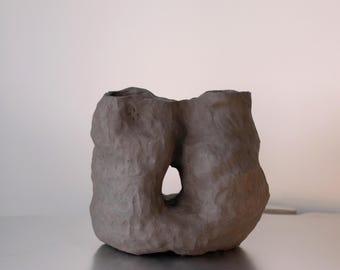 May include: A gray ceramic vase with a unique, organic shape. The vase has two openings and a hole in the center, creating a modern and abstract design.