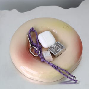 Pode incluir: Um prato de cerâmica em forma de donut com um esmalte rosa e amarelo. O prato contém um carregador sem fio branco, uma pequena caixa de metal prateada com a palavra "penco" impressa nela e uma corda roxa com um anel prateado.