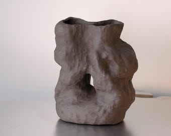 Könnte beinhalten: Eine dunkelbraune Keramikvase mit unregelmäßiger, organischer Form. Die Vase hat eine große Öffnung oben und eine kleinere Öffnung an der Seite. Die Vase steht auf einem silbernen Tisch mit einer einzigartigen, wellenförmigen Kante.