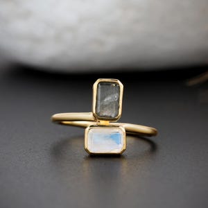 Op de afbeelding: Een gouden ring met twee rechthoekige stenen in een bypass-stijl. De bovenste steen is donkergrijs, terwijl de onderste steen een doorschijnende, iriserende witte maansteen is. De ringband is dun en gemaakt van gepolijst goud.