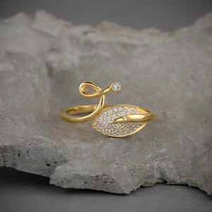Può includere: Un anello dorato con un design a foglia, incastonato con piccole pietre preziose trasparenti. L'anello presenta un dettaglio delicato e a spirale e un'unica pietra preziosa più grande. L'anello è esposto su una superficie grigia strutturata.