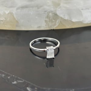 May include: A silver ring with a rectangular, clear gemstone. The ring band is thin and polished, and the gemstone is set in a simple, elegant design. The ring is displayed on a dark, reflective surface.