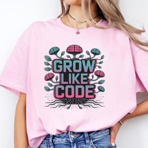 Könnte beinhalten: Hellrosa T-Shirt mit dem Schriftzug "GROW LIKE CODE" in einem farbenfrohen Design. Die Grafik zeigt gehirnförmige Elemente und eine Leiterplattenbasis. Das T-Shirt besteht aus weichem Material und eignet sich für den täglichen Gebrauch.
