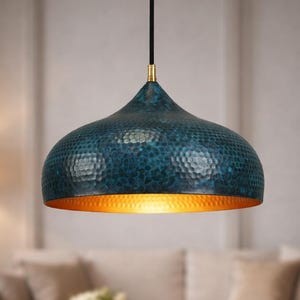 May include: A teal-colored, hammered-metal pendant light with a gold interior. The light fixture hangs from a black cord with a gold-colored accent. The dome-shaped shade has a textured surface and is suspended in a living room setting.