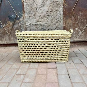 May include: A rectangular woven basket with a lid and handles. The basket is made of light-colored natural fibers and has a textured, open weave design. The basket is sitting on a brick surface.