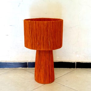 Puede incluir: Una mesa pedestal decorativa naranja. La mesa tiene una parte superior redonda y una base cilíndrica. La mesa está hecha de un material tejido y es de color naranja vibrante. La mesa es adecuada para uso en interiores o exteriores.