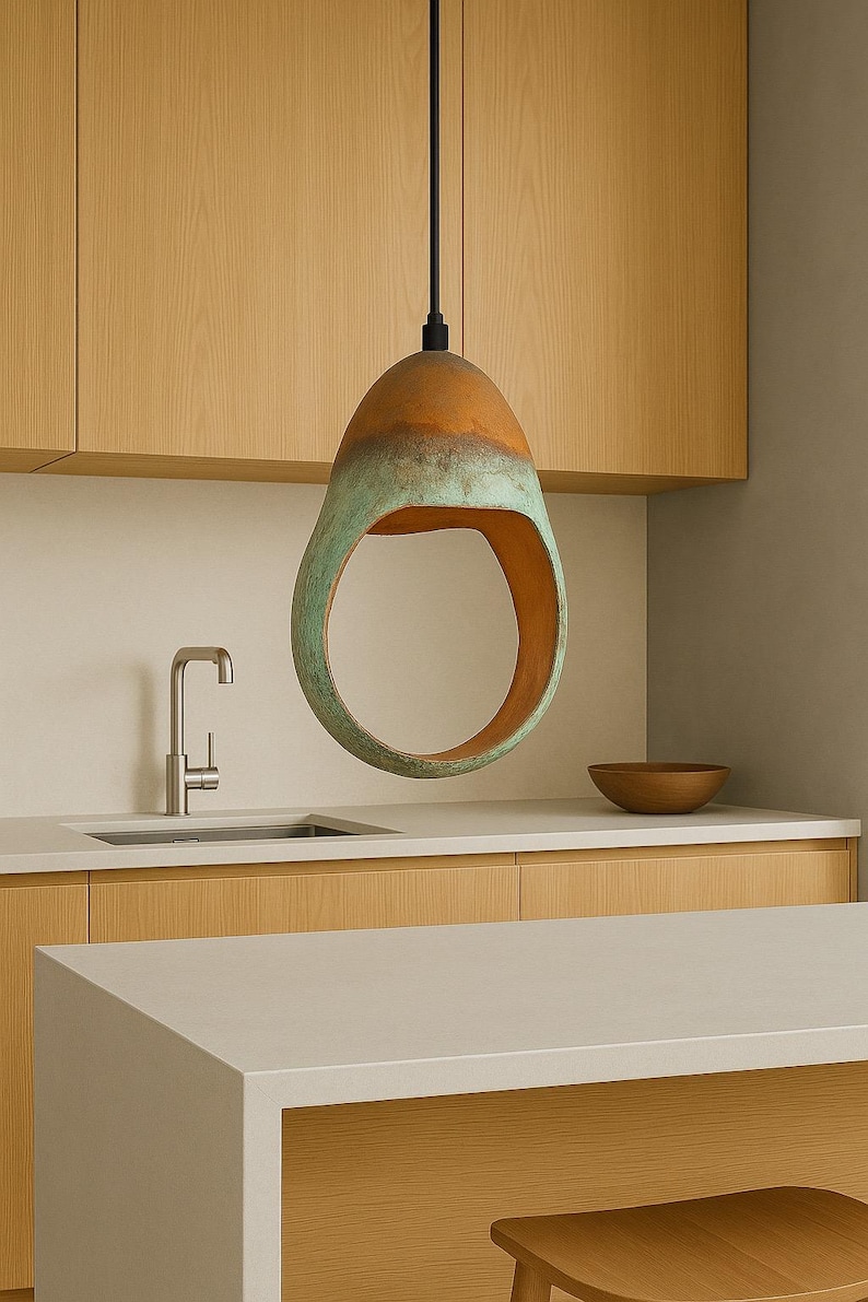 Może przedstawiać: Nowoczesna lampa wisząca o unikalnym, otwartym pierścieniowym designie. Lampa ma kształt kropli, z miedzianą g&oacute;rą i zielono-niebieskim dołem. Wisi na czarnym kablu w kuchni z jasnymi drewnianymi szafkami i kranem ze stali nierdzewnej.