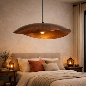Pode incluir: Uma grande luminária pendente de cor cobre pende sobre uma cama. A luminária tem um formato orgânico único com uma superfície texturizada. A luz quente da lâmpada no interior ilumina o ambiente, criando uma atmosfera acolhedora. O quarto apresenta tons neutros e almofadas decorativas.