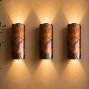 May include: Three cylindrical wall sconces with a textured, copper-colored finish. Each sconce emits a warm, downward-facing light, creating a dramatic effect. The sconces are mounted on a neutral-toned wall, with a hint of greenery in the corner.