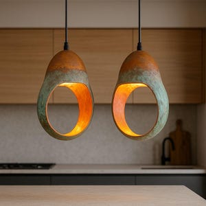 Może przedstawiać: Dwie lampy wiszące o unikalnym designie. Lampy mają kształt kropli z dużym okrągłym otworem. Zewnętrzna strona to mieszanka brązu i zieleni, a wnętrze świeci ciepłym pomarańczowym światłem. Wiszą na czarnych przewodach.