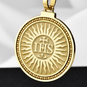 May include: Gold chain necklace with a round pendant featuring a sunburst design and the inscription "IHS" in the center.