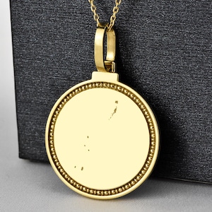 May include: A gold pendant necklace with a circular charm displaying a map design. The charm has a beaded edge and is suspended from a fine chain. The necklace is shown against a dark backdrop.