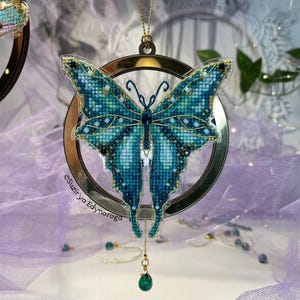以下が含まれることがあります： 金色のアクセントが付いたターコイズブルーの蝶のオーナメントが、金色のフレームが付いた透明なプラスチックの円の中に吊り下げられています。オーナメントは、緑色の宝石がついた金色のチェーンから吊り下げられています。