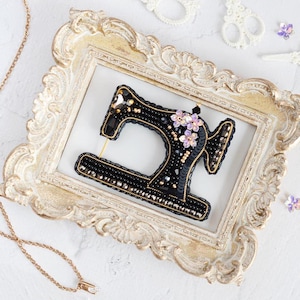 May include: A black and gold beaded sewing machine brooch with purple flowers. The brooch is displayed in a white ornate frame.