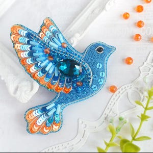 May include: A blue and orange bird brooch with a teal rhinestone. The bird is made of sequins and beads.