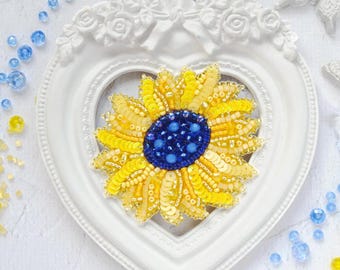 May include: A yellow and blue beaded sunflower brooch in a white heart-shaped frame. The sunflower is made of yellow and gold beads with a blue center.